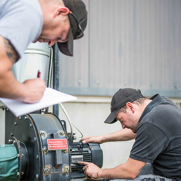 Zwei Mitarbeiter warten eine Maschine. Einer kontrolliert das Typenschild, der andere schreibt die genauen Daten in den Wartungsbericht.